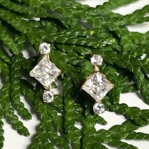 May include: A pair of gold earrings with a square-shaped diamond in the centre and two smaller diamonds on either side.