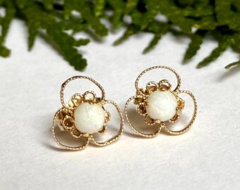 May include: A pair of gold-coloured flower-shaped stud earrings. Each earring features a white, round centre stone surrounded by a delicate, openwork design. The earrings are set against a white surface with green foliage in the background.