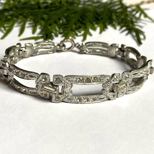 Puede incluir: Un brazalete plateado con un diseño geométrico. El brazalete presenta eslabones rectangulares y ovalados, cada uno adornado con pequeñas piedras preciosas transparentes. El cierre es un simple cierre de gancho y ojo. El brazalete se exhibe sobre una superficie blanca con follaje verde en el fondo.