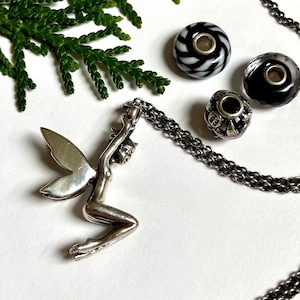 Puede incluir: Un collar con colgante de hada de plata con una cadena, acompañado de tres cuentas decorativas en blanco y negro. El collar está sobre un fondo blanco con una ramita de follaje verde.