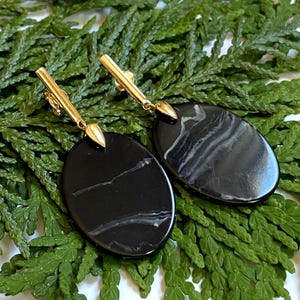 Pode incluir: Um par de brincos dourados com pingentes ovais pretos com marmoreio branco. Os brincos são exibidos sobre uma cama de folhagem verde, mostrando seu design elegante e cores contrastantes.