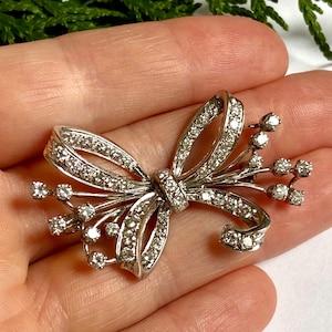 May include: A silver brooch in the form of a bow, adorned with many small, glittering gemstones. The bow design features intricate curves and a central knot, with gemstone sprigs extending from the sides, creating a floral effect.