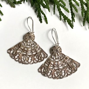 Puede incluir: Un par de pendientes de filigrana plateada con un diseño en forma de abanico. Los pendientes son delicados e intrincados, con un patrón floral.