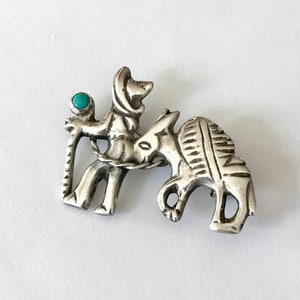 Puede incluir: Broche de metal plateado que representa una figura con sombrero montando un burro. La figura sostiene un bastón con una piedra turquesa. El burro tiene marcas grabadas. La broche es de estilo suroeste.