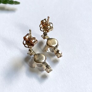 May include: A pair of gold earrings with clear gemstones. The earrings have a dangling design with a small gemstone at the top and a larger gemstone at the bottom.
