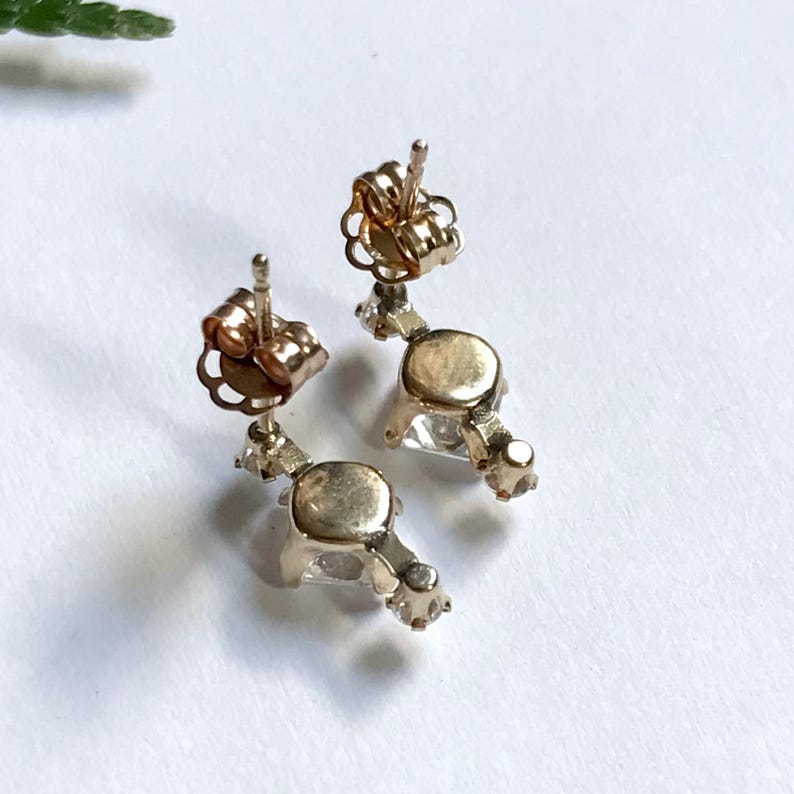 May include: A pair of gold earrings with clear gemstones. The earrings have a dangling design with a small gemstone at the top and a larger gemstone at the bottom.