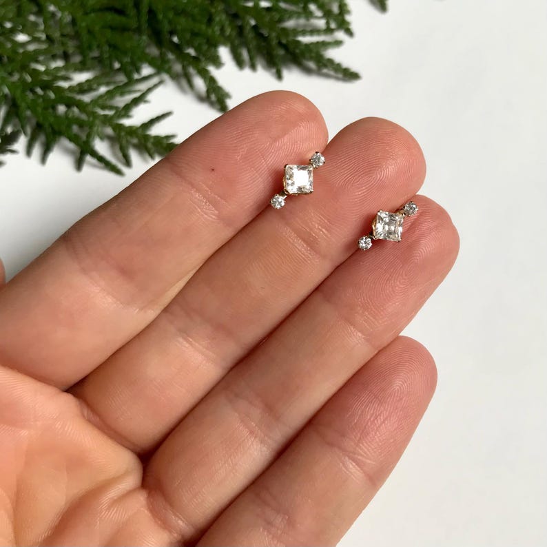 May include: A pair of gold earrings featuring small square-shaped clear gemstones. Each earring has a larger square gemstone in the centre and two smaller round gemstones on either side.