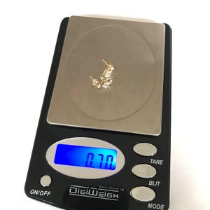 May include: A digital scale with a blue display showing a weight of 0.70 grams. A pair of gold earrings with small clear stones are on the scale.
