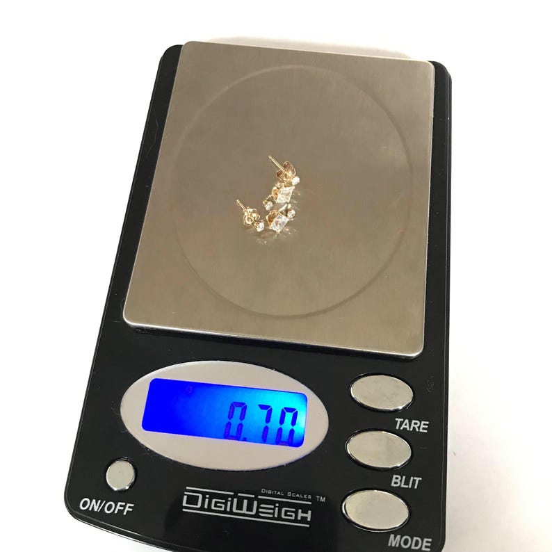 May include: A digital scale with a blue display showing a weight of 0.70 grams. A pair of gold earrings with small clear stones are on the scale.