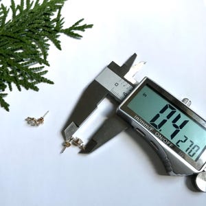 May include: Close-up of a silver digital caliper measuring a small gold earring with clear stones. The caliper reads 0.21 inches.