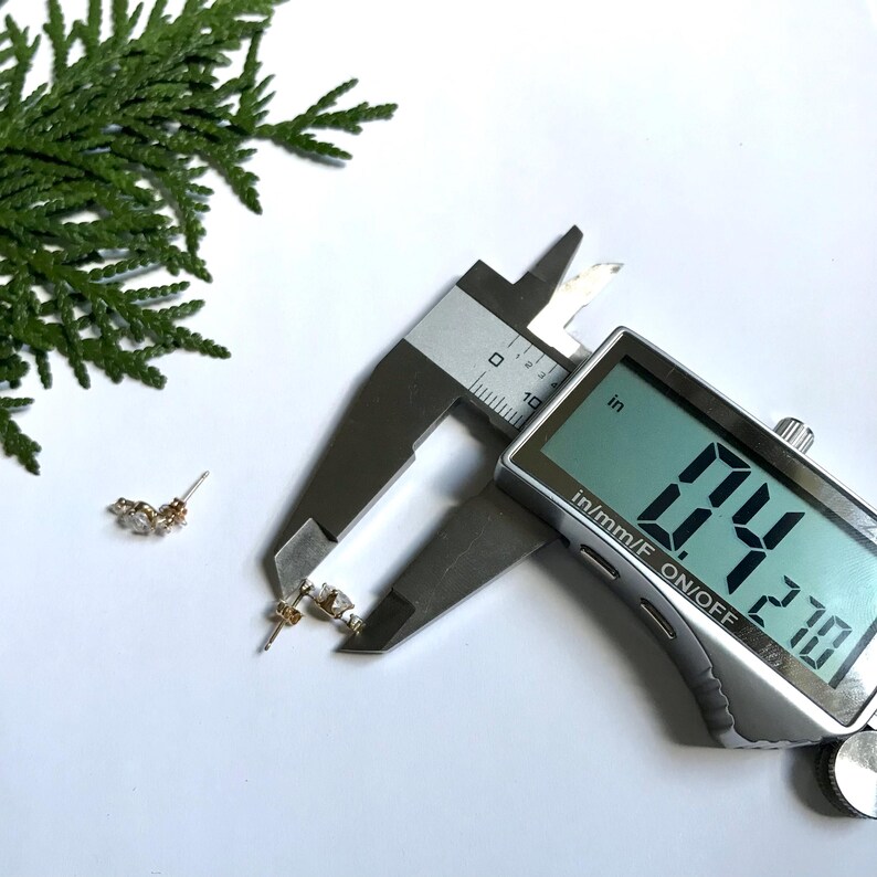 May include: Close-up of a silver digital caliper measuring a small gold earring with clear stones. The caliper reads 0.21 inches.