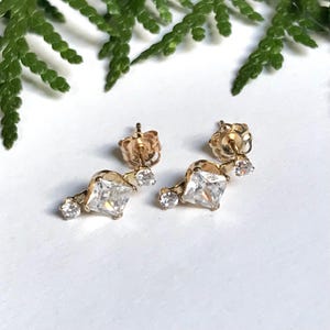May include: A pair of gold earrings featuring a square clear stone and smaller clear stones. The earrings have a delicate, minimalist design.