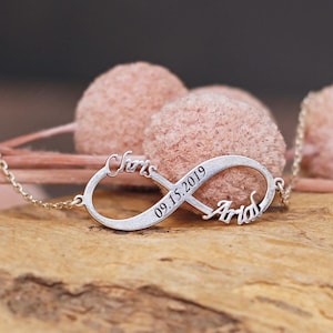 Könnte beinhalten: Silberne Unendlichkeitskette mit den Namen "Chris" und "Arid" sowie dem Datum "09.15.2019" eingraviert. Die Kette liegt auf einer Holzoberfläche mit rosa Dekorationskugeln im Hintergrund.
