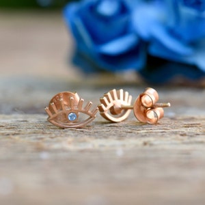 Könnte beinhalten: Ein Paar goldfarbene Ohrringe mit einem blauen Augen-Design. Die Ohrringe sind klein und zart und wären ein tolles Geschenk für eine besondere Person.