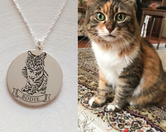 Può includere: Una collana d'argento con un ciondolo rotondo che presenta un'immagine incisa in bianco e nero di un gatto. Il ciondolo ha la parola "Bodie" incisa su uno stendardo sotto l'immagine.