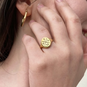 May include: Gold signet ring with a circular design featuring the letters "T", "R", "S", and "M" engraved in a cross pattern.