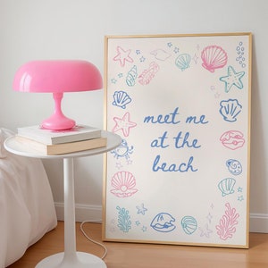May include: A pink lamp with a white base sits on a white side table next to a bed. A framed print with a gold frame hangs on the wall behind the table. The print features a white background with pink and blue illustrations of seashells, starfish, and seaweed. The text "meet me at the beach" is written in blue on the print.