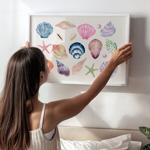 May include: A framed watercolor painting of various seashells in shades of pink, blue, purple, and green. The shells are arranged in a random pattern on a white background.