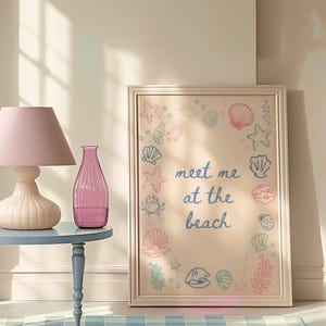 May include: A framed print with a light beige background and a border. The print features a circle of sea-themed illustrations, including seashells, starfish, and seaweed. The text "meet me at the beach" is written in blue and pink lettering in the centre of the print.