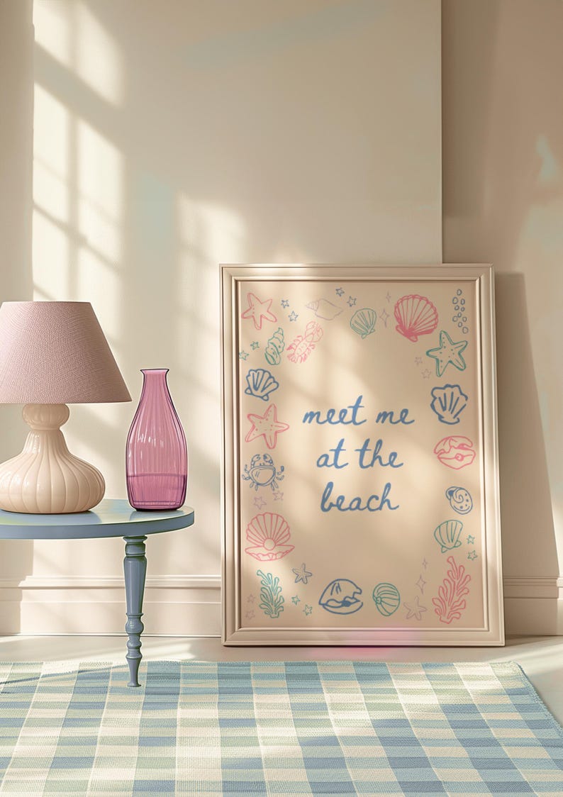 May include: A framed print with a light beige background and a border. The print features a circle of sea-themed illustrations, including seashells, starfish, and seaweed. The text "meet me at the beach" is written in blue and pink lettering in the centre of the print.