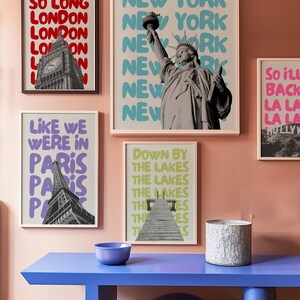 May include: A collection of five framed prints with different cityscapes and slogans. The prints are hung on a light pink wall. The prints feature the Eiffel Tower, Big Ben, the Statue of Liberty, and a pier. The slogans on the prints include "So Long London", "Like We Were in Paris", "Welcome to New York", "Down by the Lakes", and "So I'll Back La La La".