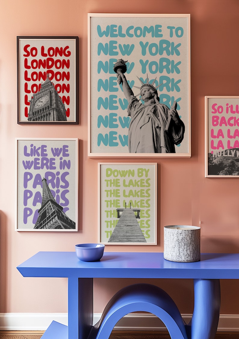 May include: A collection of five framed prints with different cityscapes and slogans. The prints are hung on a light pink wall. The prints feature the Eiffel Tower, Big Ben, the Statue of Liberty, and a pier. The slogans on the prints include "So Long London", "Like We Were in Paris", "Welcome to New York", "Down by the Lakes", and "So I'll Back La La La".