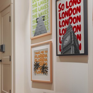 May include: Three framed art prints on a white wall. The print on the left has a green background with the text "Down by the Lakes" repeated in a stacked pattern. The print in the middle has a beige background with the text "Take me to Florida" repeated in a stacked pattern. The print on the right has a black frame and a black and white photo of Big Ben with the text "So Long London" repeated in a stacked pattern in red.