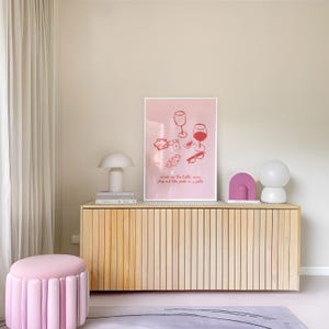 May include: A pink and white framed illustration of two wine glasses, playing cards, and glasses on a table with the text "cards on the table, wine flowing and the faith in a fella". The illustration is hanging on a wall above a light wood cabinet with two lamps on the top.
