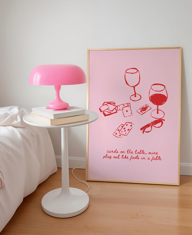 May include: A pink and gold framed art print with a pink background and red illustrations of two wine glasses, playing cards, and sunglasses. The text on the print reads "cards on the table, mine play out like fools in a fable".