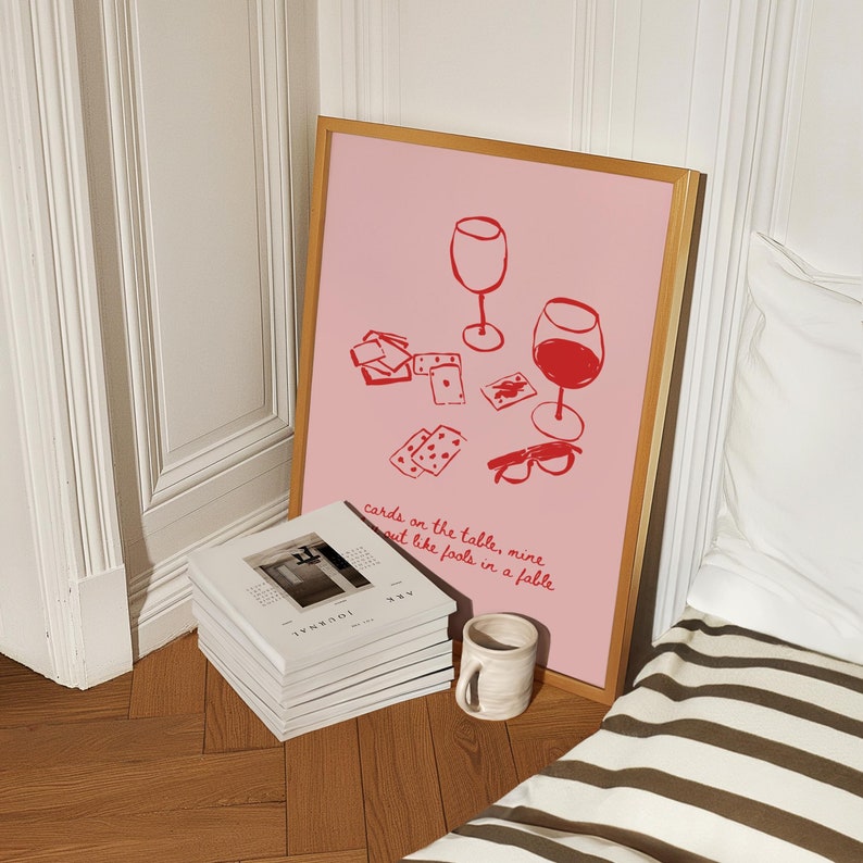 May include: A framed pink poster with a red illustration of two wine glasses and playing cards. The text on the poster reads "cards on the table, mine out like fools in a fable".