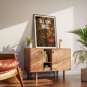May include: A framed poster with the text "ALL TOO WELL" in white letters on a background of a forest scene in shades of brown, orange, and yellow. The poster is hanging on a wall above a wooden cabinet with a hand sculpture on the right side.