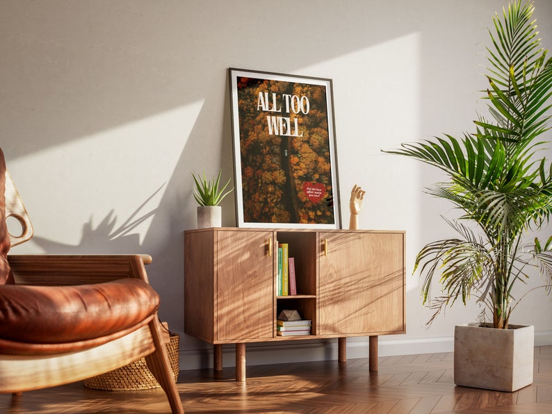 May include: A framed poster with the text "ALL TOO WELL" in white letters on a background of a forest scene in shades of brown, orange, and yellow. The poster is hanging on a wall above a wooden cabinet with a hand sculpture on the right side.