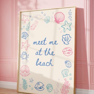 May include: A beige poster with a gold frame features a blue and pink illustration of seashells, starfish, and seaweed. The text "meet me at the beach" is written in blue cursive.