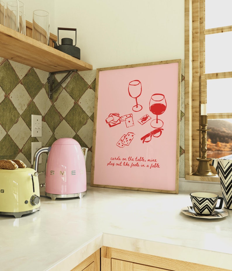 May include: A pink kitchen counter with a pink kettle, a yellow toaster, and a framed print with a pink background and red illustrations of wine glasses, playing cards, and glasses. The print has the text "cards on the table, mine play out like fools in a fable".