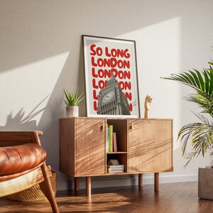 May include: A framed print with a black frame and a white background. The print features a photo of Big Ben in London with the text "So Long London" repeated in red letters.