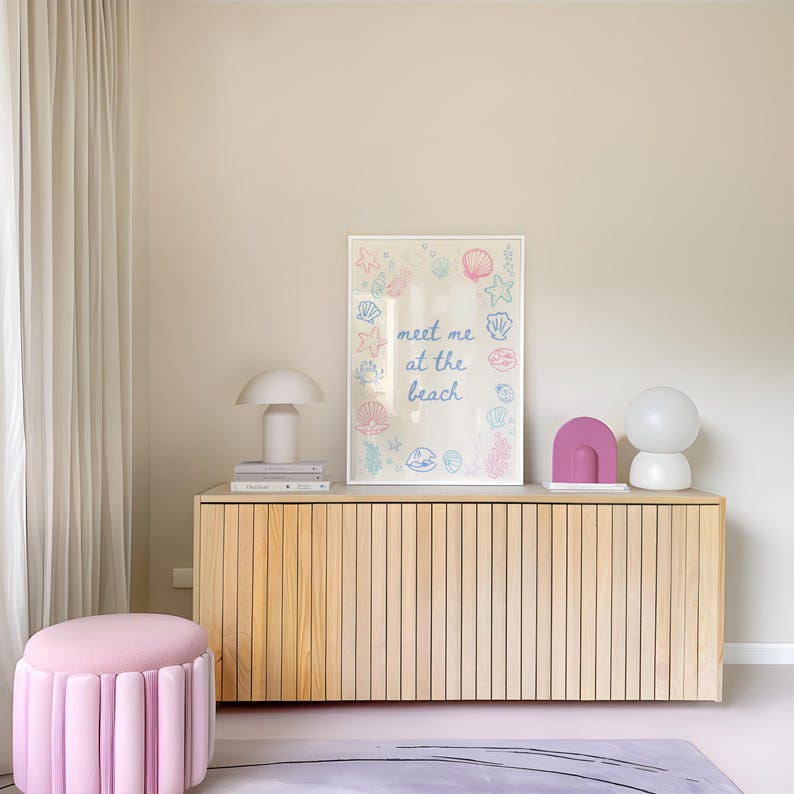 May include: A light wood cabinet with a framed print that says "meet me at the beach" surrounded by illustrations of seashells and starfish. There are two lamps and a pink decorative object on top of the cabinet.