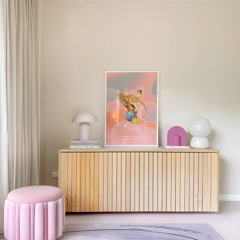 May include: A wooden cabinet with a light wood finish and vertical slats. The cabinet has two doors and is decorated with a framed print of a woman riding a tiger on a ball, two lamps, and a stack of books. The print has a pink and gray background with the text "I am stronger, I am braver, I am more powerful than I ever thought I could be."