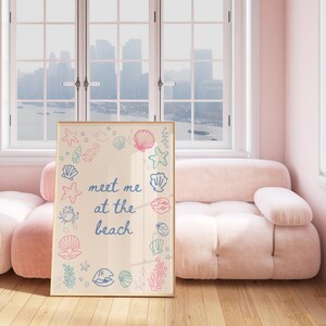 May include: A framed print with a light beige background and a blue inscription that reads "meet me at the beach". The inscription is surrounded by a border of colourful illustrations of seashells, starfish, and seaweed.