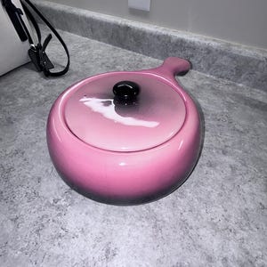 Puede incluir: Una olla de fondue redonda de cerámica rosa con tapa a juego y un solo mango. La olla presenta un acabado brillante que se oscurece hacia la base. Un pequeño pomo negro redondo está centrado en la tapa, la cual tiene una superficie lisa y reflectante.