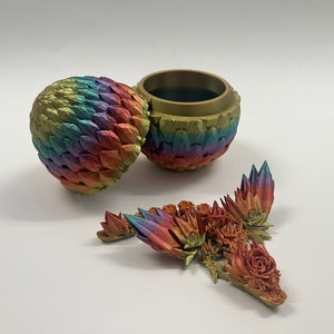 May include: A rainbow-colored dragon egg-shaped trinket box with a removable lid. The box is open and the lid is laying next to it. There are several small, colorful dragon scales scattered around the box.