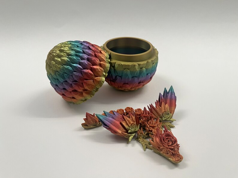 May include: A rainbow-colored dragon egg-shaped trinket box with a removable lid. The box is open and the lid is laying next to it. There are several small, colorful dragon scales scattered around the box.