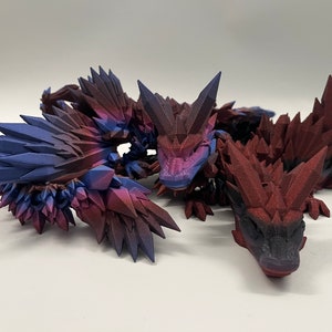 May include: Three 3D printed dragon figurines with a crystal-like texture. The dragons are red, purple, and blue with a black base.