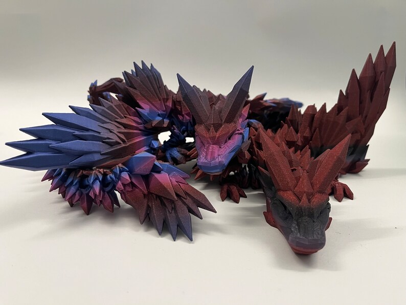 May include: Three 3D printed dragon figurines with a crystal-like texture. The dragons are red, purple, and blue with a black base.