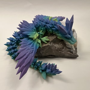 May include: A 3D printed crystal dragon figurine with a gradient of blue, green, and purple colors. The dragon is perched on a gray rock.