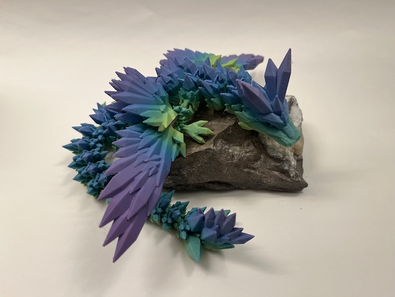 May include: A 3D printed crystal dragon figurine with a gradient of blue, green, and purple colors. The dragon is perched on a gray rock.