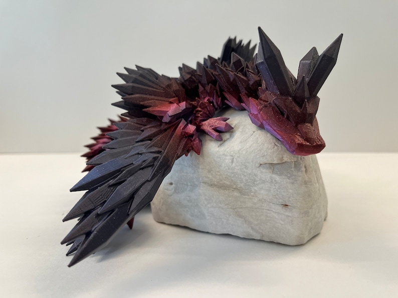 May include: A 3D printed dragon figurine with a black and red crystal-like design. The dragon is lying down on a white rock.