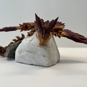 May include: A wooden sculpture of a dragon with brown and red scales, resting on a white rock.