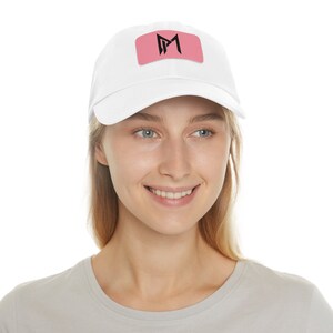 May include: A white baseball cap with a pink patch featuring a black stylized letter "M" on the front.