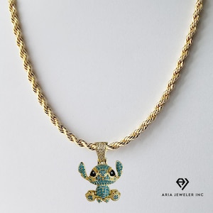 May include: A gold-colored rope chain necklace with a pendant featuring a cartoon character. The pendant is adorned with blue and clear gemstones, with black accents. The necklace is displayed against a white background.