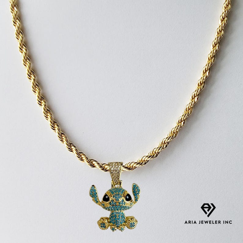 May include: A gold-colored rope chain necklace with a pendant featuring a cartoon character. The pendant is adorned with blue and clear gemstones, with black accents. The necklace is displayed against a white background.
